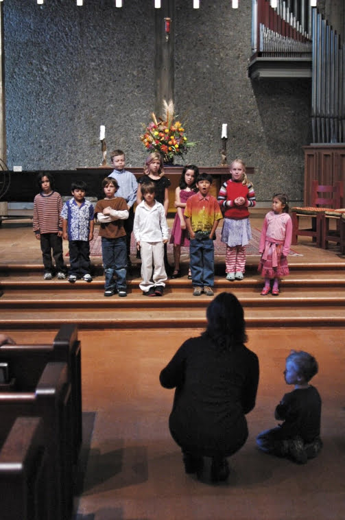 10 young singers standing on the chancel steps, singing; MIchèle kneeling in front of them, back to the camera, conducting; a four-year old looks on, supervising.